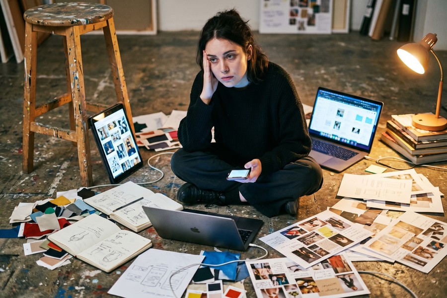 Freelance designer at desk with multiple messaging apps open across laptop and phone screens